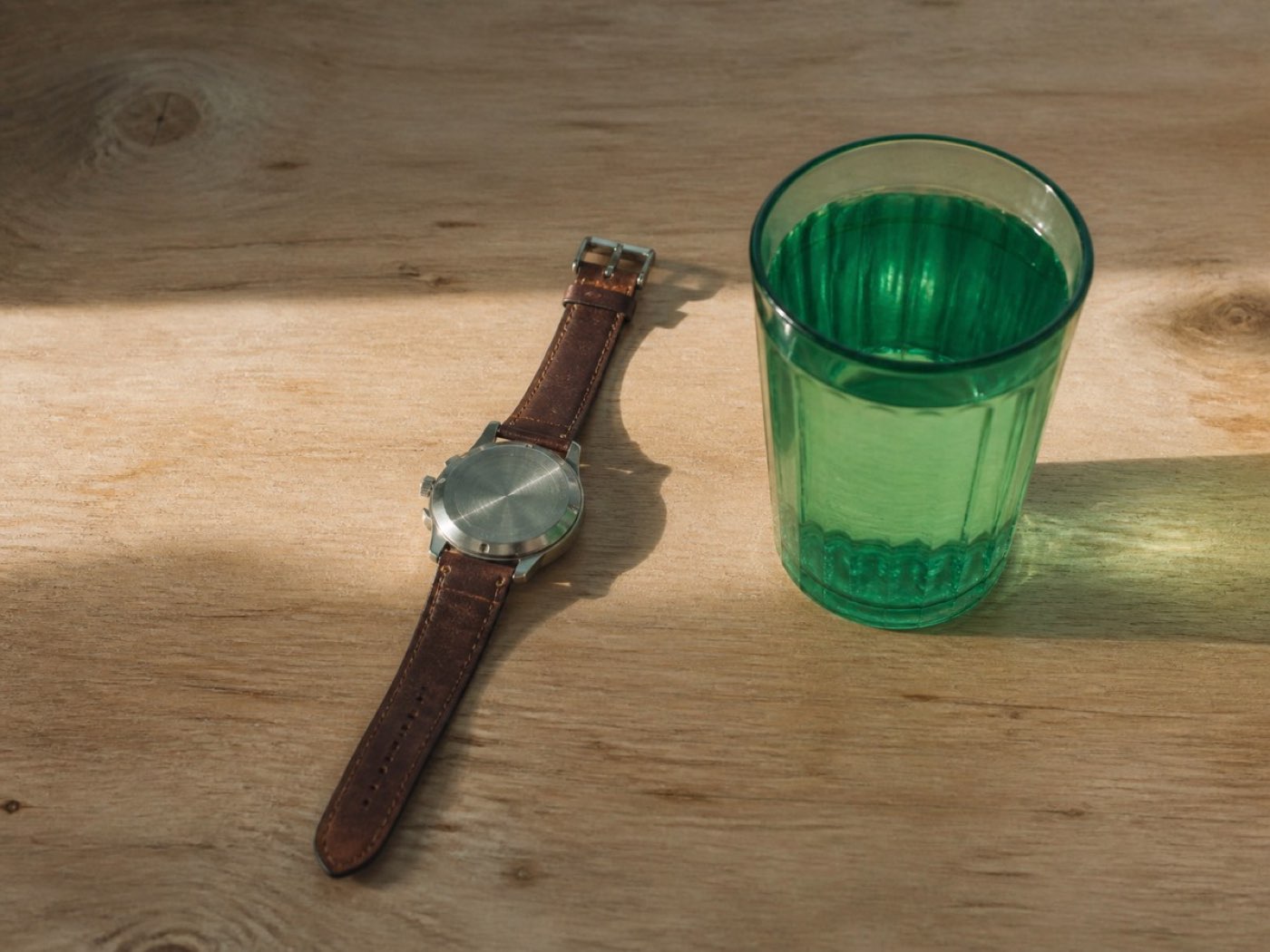 A wristwatch beside a filled glass on a desk in early morning light