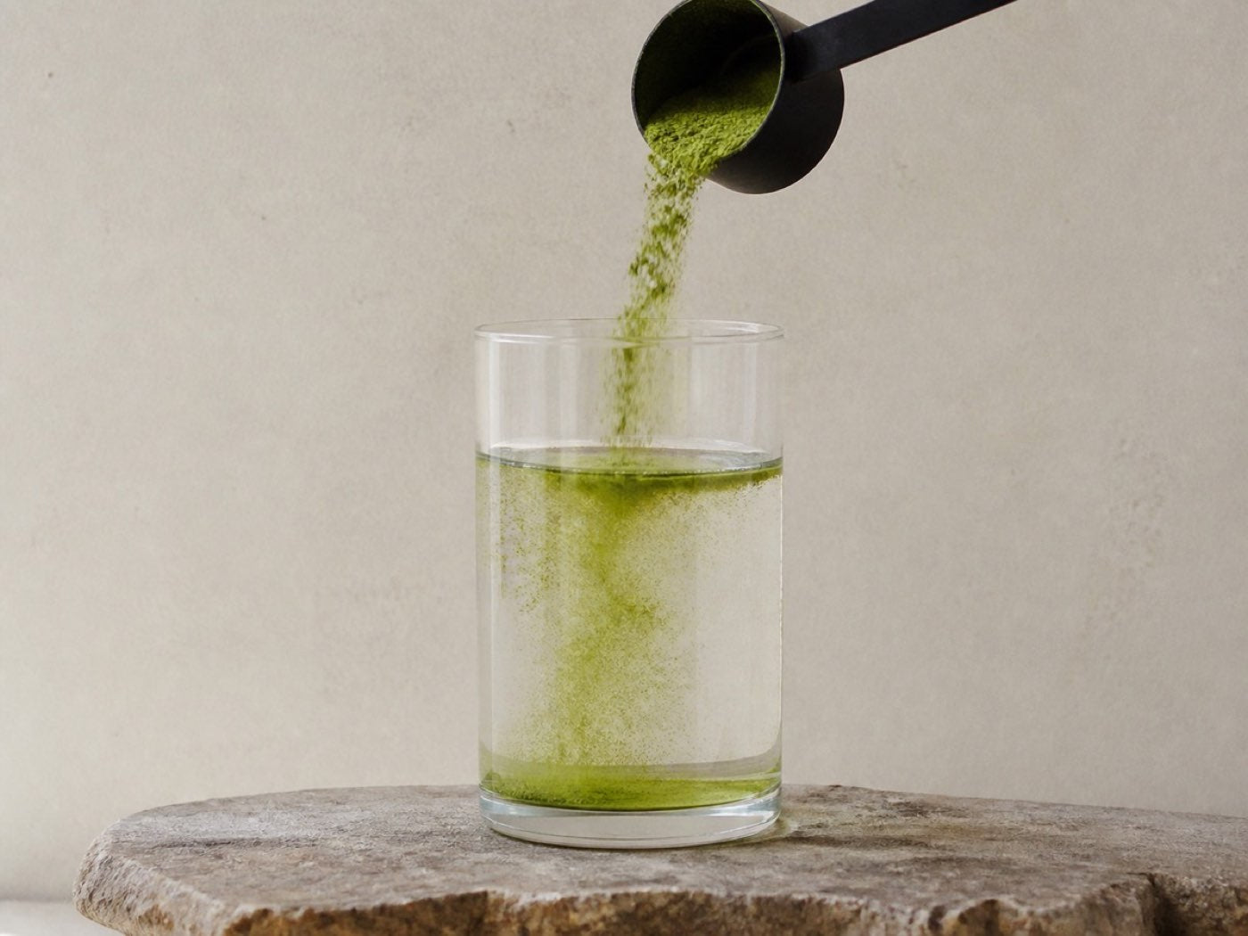 A close view of green powder falling from a black scoop into a glass of water